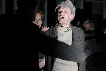 Turkishattuun ja -takkiin pukeutunut mies seisoo dramaattisessa valaistuksessa ja vaikuttaa ahdistuneelta, suu auki kuin huutaisi. Hänen vierellään on muita ihmisiä, joista osa näkyy osittain, pimeässä, jännittyneessä ympäristössä.