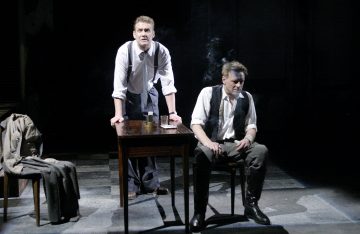 Kaksi miestä, joilla on rullattu paita ja henkselit, esittää näyttämöllä dramaattisen kohtauksen; toinen seisoo pöydän takana eteenpäin nojautuneena, kun taas toinen istuu savukkeen kanssa alaspäin katsoen. Takki on ripustettu heidän vieressään olevan tuolin päälle.