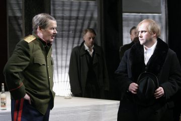 Kaksi aikakauden pukuihin pukeutunutta miestä seisoo näyttämöllä vastakkain; toisella on sotilasunivormu, toisella musta takki ja hattu. Taustalla seisoo kaksi muuta miestä. Pullo ja lasi ovat läheisellä pöydällä.