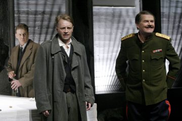 Kolme miestä seisoo sisätiloissa; yksi tummapukuinen mies vaikuttaa ahdistuneelta, toinen sotilaspukuinen mies hymyilee itsevarmasti, ja kolmas pukuinen mies seisoo taustalla lasia kädessään, ja heidän takanaan on valkoinen pöytä ja harmaa tausta.