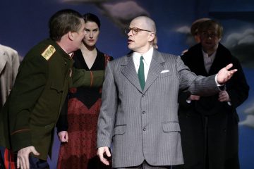 Sotilasunivormuun pukeutunut mies osoittaa eleellä kohti kaljua miestä, jolla on harmaa puku ja silmälasit. Kaksi muuta henkilöä, punamekkoon pukeutunut nainen ja tummaan takkiin pukeutunut mies, seisovat taustalla lavasteissa.