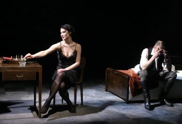 Mustapukuinen nainen istuu pöydän ääressä tupakoimassa, kun taas mies istuu taustalla sängyllä pitäen päätään käsissään. Kohtaus on hämärästi valaistu ja dramaattinen, mikä viittaa hahmojen väliseen jännitteeseen.