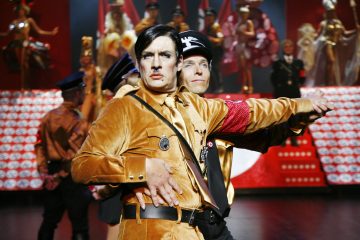 Kaksi esiintyjää natsiunivormuja muistuttavissa teatteripuvuissa tanssii lavalla dramaattisin ilmein. Taustalla näkyvät kirkkaanpunaiset ja kultaiset lavasteet ja muut pukuun pukeutuneet näyttelijät.