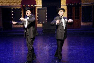 Kaksi esiintyjää smokissa ja rusettihatuissa tanssii lavalla, elehtii kepillä ja hymyilee kirkkaiden näyttämövalojen alla, ja taustalla on teatteri, jossa on teltan valot ja ovet.