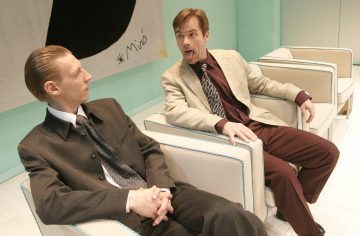 Kaksi pukumiestä istuu nojatuoleissa; toinen näyttää vakavalta kädet ristissä, kun taas toinen tekee leikkisän ilmeen ja työntää kielensä ulos ensimmäisen miehen suuntaan. Ympäristö näyttää olevan moderni, vaalea huone.