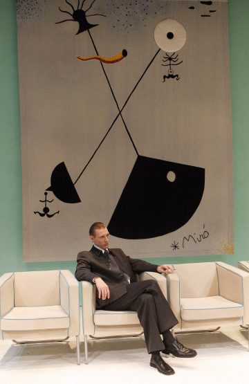 Tummapukuinen mies istuu kermanvärisessä nojatuolissa, jonka vieressä on lisää tuoleja. Hänen takanaan roikkuu vaaleaa seinää vasten suuri geometrisia muotoja ja viivoja esittävä abstrakti taideteos, jonka on signeerannut Miró.