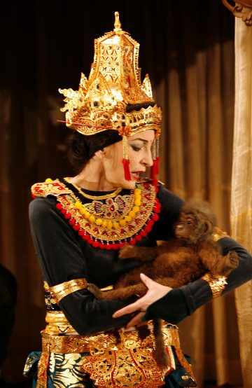 Balilainen tanssija, jolla on koristeellinen perinteinen balilainen puku ja kultainen päähine, pitelee lavalla pientä ruskeaa apinaa, ja taustalla on runsaat kultaiset verhot.