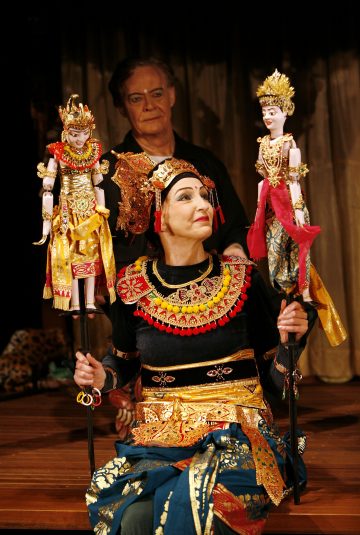 Koristeelliseen perinteiseen pukuun pukeutunut esiintyjä pitää kahta taidokkaasti pukeutunutta nukkea keppien varassa, kun toinen henkilö seisoo heidän takanaan puisella näyttämöllä, jonka taustalla on verho.