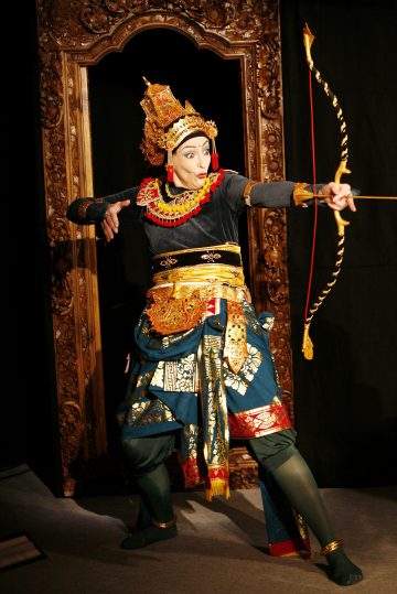 Koristeelliseen perinteiseen indonesialaiseen asuun pukeutunut henkilö poseeraa kultaisen jousen ja nuolen kanssa, seisoo taidokkaasti veistetyn puukehyksen edessä ja tekee dramaattisen tanssieleen.