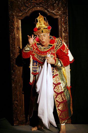 Esiintyjä, jolla on taidokas, värikäs puku ja kultainen kruunu, seisoo paljain jaloin, valkoinen liina kädessään ja dramaattinen ilme, ja häntä kehystää koristeellinen puurakenne tummalla taustalla.