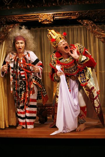 Kaksi näyttelijää näyttämöllä taidokkaissa puvuissa: toisella on villi harmaa tukka ja raidallinen asu, toisella koristeellinen punainen ja kultainen asu, molemmat tekevät dramaattisia ilmeitä verhotaustaa vasten.