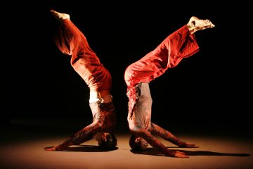 Kaksi punaisiin pukuihin pukeutunutta akrobaattia tasapainoilee päänsä ja käsiensä varassa, vartalo kaaressa ja jalat ylöspäin ojennettuina, luoden peilimäisen, symmetrisen poseerauksen tummaa taustaa vasten.