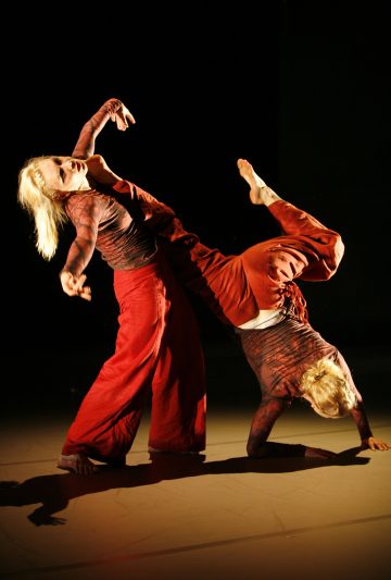 Kaksi vaaleatukkaista tanssijaa, joilla on punaiset housut ja kuviolliset topit, esittävät ilmeikkään rutiinin tummalla taustalla. Toinen seisoo pystyasennossa kädet kohotettuna, kun taas toinen on ylösalaisin tasapainoillen yhden käden varassa.