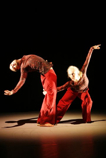 Lavalla esiintyy kaksi tanssijaa, joilla on samanlaiset punaiset asut, joista toinen kaartuu dramaattisesti taaksepäin, kun taas toinen nojaa eteenpäin ojennettu käsi, ja molemmat ovat valonheittimen valaisemia tummalla taustalla.