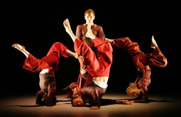 Neljä punaisiin asuihin pukeutunutta tanssijaa tekee akrobaattisia poseerauksia lattialla, tasapainoilee hartioillaan ja päillään, kun yksi henkilö seisoo pystyasennossa heidän takanaan pimeällä näyttämöllä.