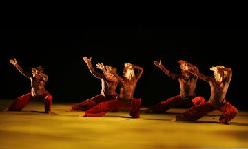 Viisi tanssijaa punaisissa housuissa ja tummissa topeissa esittää lavalla synkronoidun rutiinin, joka on ilmeikäs, polvet koukussa ja kädet kohotettuna, lämpimässä, dramaattisessa valaistuksessa tummaa taustaa vasten.