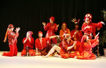 Ryhmä ihmisiä, joilla on elinvoimaiset punaiset puvut ja hatut, poseeraa ja elehtii vilkkaasti lavalla, osa istuen ja osa seisten, tummaa taustaa vasten, jonka oikealla puolella on ruukkukasvi.