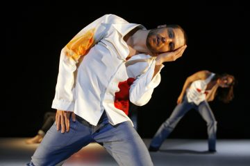 Valkoiseen paitaan ja farkkuihin pukeutunut henkilö nojautuu sivuttain ja nojaa päänsä käteensä, ja taustalla on muita samankaltaisissa asennoissa olevia henkilöitä, jotka kaikki esiintyvät lavalla, jolla on tumma tausta.