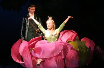 Satuprinsessaksi pukeutunut nainen istuu lavalla suuren, vaaleanpunaisen kukan sisällä hymyillen ja kädet kohotettuna. Hänen takanaan seisoo vihreä puku ja kruunu päällään mies, jota valaisee näyttämövalaistus.
