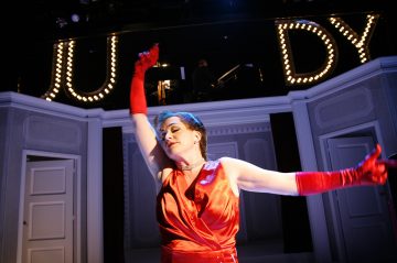 Punamekkoon ja pitkiin punaisiin hanskoihin pukeutunut esiintyjä poseeraa dramaattisesti lavalla JUDYn valaiseman kyltin alla. Lavan tausta on valkoinen ovineen ja pylväineen.