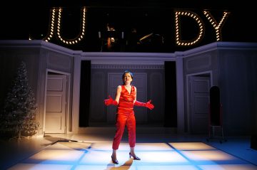 Punaiseen asuun ja hanskoihin pukeutunut esiintyjä laulaa intohimoisesti kirkkaasti valaistulla lavalla, vasemmalla joulukuusi ja yläpuolella valaistuin kirjaimin kirjoitettu JUDY. Taustalla soittavat muusikot.
