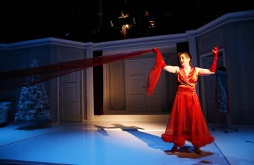 Kirkkaanpunaiseen mekkoon ja hanskoihin pukeutunut henkilö pitelee dramaattisesti pitkää punaista huivia lavalla, jossa on talvikoristeita, kuten valkoinen joulukuusi, ja taustalla soittaa kaksi muusikkoa.