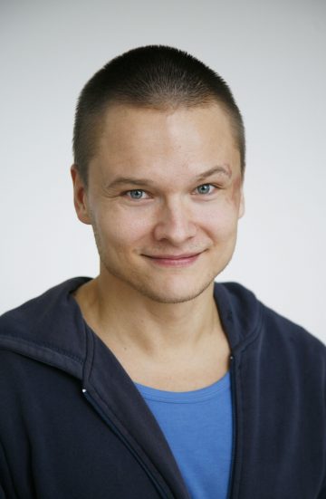 Hymyilevä lyhythiuksinen henkilö, jolla on sininen paita ja tumma vetoketjullinen huppari, poseeraa vaaleaa taustaa vasten.
