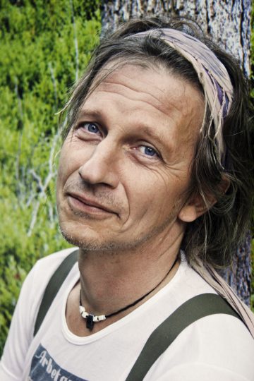 Keski-ikäinen henkilö, jolla on siniset silmät ja sekaiset hiukset, päässään hiuspanta, kaulakoru ja valkoinen paita, nojaa puuta vasten, jonka taustalla on vehreyttä, ja katsoo rauhallisesti kameraan.