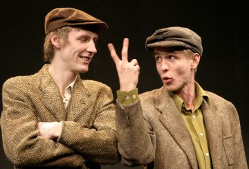 Kaksi ruskeisiin tweed-takeihin ja litteisiin lippiksiin pukeutunutta miestä seisoo mustaa taustaa vasten. Toinen mies on kädet ristissä ja hymyilee, kun taas toinen pitää kahta sormea ylhäällä ja puhuu vilkkaasti.