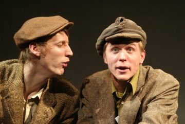 Kaksi vanhanaikaiseen, ruskeaan tweed-takkiin ja litteisiin lippiksiin pukeutunutta miestä istuu tiiviisti vierekkäin. Toinen mies katsoo toista, joka tuijottaa hämmästyneenä eteenpäin. Tausta on tumma ja yksinkertainen.