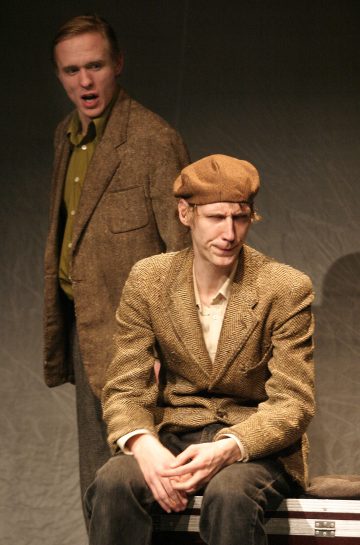 Kaksi ruskeatakkista miestä esiintyy lavalla; toinen istuu ja hänellä on litteä lippalakki, kun taas toinen seisoo hänen takanaan ilmeikkäät kasvot dramaattiseen kohtaukseen viittaavasti.