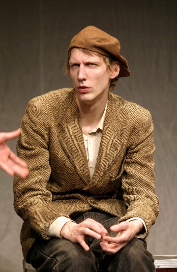 Henkilö, jolla on ruskea baretti ja ruskea kalanruototakki, istuu hämmentyneenä kädet ristissä. Toinen henkilö ojentaa kätensä heitä kohti kuvan vasemmalta puolelta.