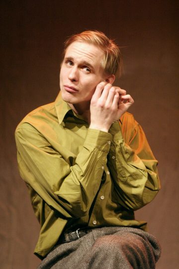 Vaaleahiuksinen henkilö, jolla on vihreä paita ja harmaat housut, istuu laittaen korvakorua ja katselee mietteliäästi ylöspäin pehmeästi valaistua ruskeaa taustaa vasten.