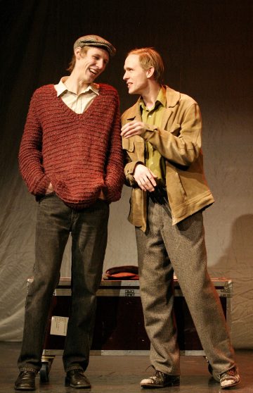 Kaksi miestä seisoo lavalla hymyillen ja keskustellen. Toisella on punainen pusero, kauluspaita, litteä lakki ja tummat housut. Toisella on ruskehtava takki, vihreä paita ja harmaat housut. Taustalla on nähtävissä matka-arkku ja verho.