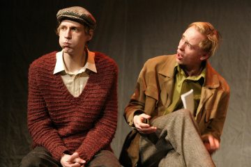 Kaksi miestä istuu lavalla. Vasemmalla istuva mies, jolla on punainen villapaita ja lippis, paiskoo poskia ja katsoo muualle. Oikeanpuoleinen mies, jolla on ruskea takki ja vihreä paita, näyttää puhuvan hänelle vilkkaasti.