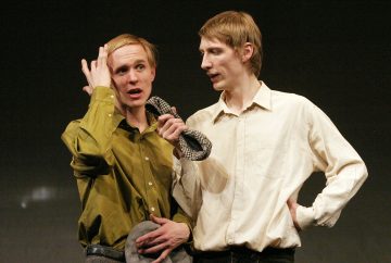 Kaksi miestä seisoo yhdessä lavalla; toinen vihreään paitaan pukeutuneista elehtii kädellään päänsä lähellä ja pitelee harmaata hattua, kun taas toinen valkoiseen paitaan pukeutunut mies pitää mikrofonia itseään kohti.