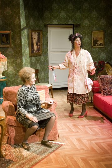 Kaksi naista vintage-tyylisessä olohuoneessa; toinen istuu nojatuolissa leopardikuvioinen asu yllään, kun taas toinen seisoo verisessä yöpaidassaan veitsi ja hattu kädessään ja näyttää kiihtyneeltä. Asetelma vaikuttaa teatraaliselta.