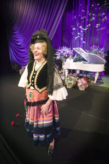 Värikkääseen kansallispukuun ja kiharaiseen peruukkiin pukeutunut esiintyjä seisoo lavalla hymyillen, kun pianisti soittaa valkoista flyygeliä kukkien ja violettien verhojen ympäröimänä taustalla.