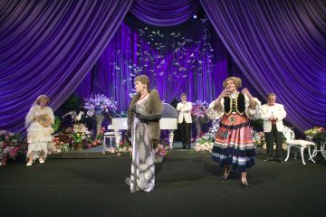 Ryhmä hienostuneisiin pukuihin pukeutuneita ihmisiä esiintyy lavalla, joka on koristeltu violeteilla verhoilla, kukilla ja valkoisella pianolla; edessä seisoo kaksi naista, toinen hopeisessa puvussa ja toinen värikkäässä kansallispuvussa.