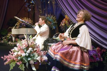 Valkoiseen smokkiin pukeutunut mies soittaa valkoista pianoa kukkien täyttämällä näyttämöllä, kun värikkääseen perinteiseen pukuun pukeutunut hymyilevä esiintyjä istuu vieressä lasi kädessään violetin kankaan ja kukkakoristeiden alla.