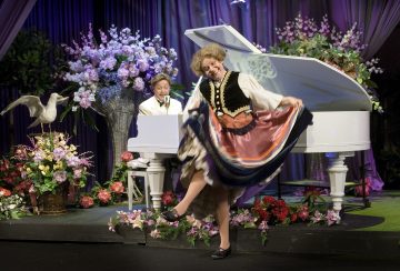 Värikkääseen kansallispukuun pukeutunut esiintyjä tanssii iloisesti valkoisen flyygelin edessä, kun valkoiseen takkiin pukeutunut henkilö soittaa ja laulaa. Lava on koristeltu värikkäillä kukilla ja violeteilla verhoilla.