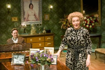 Kuvioituun mekkoon pukeutunut nainen seisoo ja puhuu vintage-huoneessa, kun mies istuu pianon takana. Huoneessa on vihreä tapetti, kehystettyjä valokuvia, kukkia ja suuri naisen muotokuva seinällä.