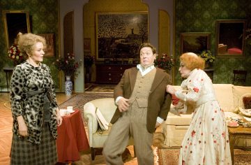 Kolme näyttelijää näyttämöllä esittää dramaattisen kohtauksen vintage-olohuoneen lavasteissa; yksi nainen vetää miestä käsivarresta, kun toinen kuviotakkiasuinen nainen katselee, ja kaikilla on liioiteltuja ilmeitä.