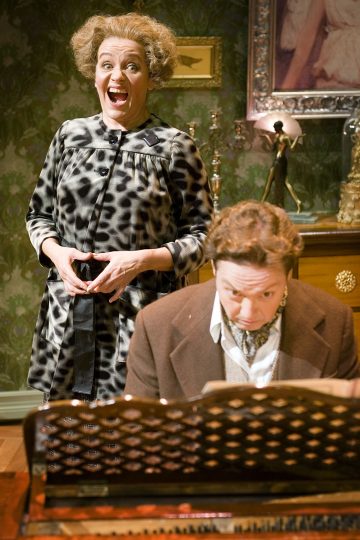 Kihara tukkainen nainen seisoo hymyillen leveästi kuviomekossaan, kun taas pukuinen mies istuu pianon ääressä ja katsoo nuotteja; he ovat tapetilla ja taideteoksilla koristellussa huoneessa.