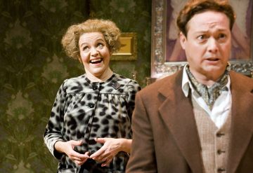 Nainen, jolla on kiharat hiukset ja kuviollinen mekko, hymyilee leveästi, kädet ristissä, kun taas ruskeaan pukuun ja kravattiin pukeutunut mies seisoo etualalla huolestuneen näköisenä. Kohtaus näyttää olevan teatteriesityksestä.