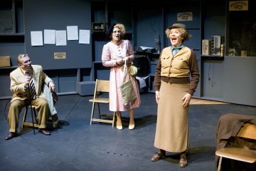 Lavalla esiintyy kolme näyttelijää. Yksi istuu puvussa nauraen, toinen seisoo vaaleanpunaisessa mekossa laukku kädessään ja kolmas hymyilee hattu päässä ja ruskeassa asussa. Lavastuksessa on tuoleja ja toimiston rekvisiittaa, jonka seinällä on papereita.