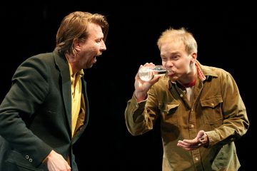 Lavalla on kaksi miestä, joista toinen tummaan takkiin ja keltaiseen paitaan pukeutunut näyttää puhuvan tai huutavan, kun taas toinen, ruskeaan takkiin pukeutunut mies juo vesilasillista ja näyttää huvittuneelta. Tausta on musta.