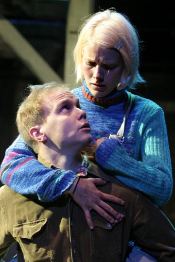 Nuori nainen, jolla on lyhyet vaaleat hiukset, katsoo vakavana alaspäin nuorta miestä syleillen häntä takaapäin. Mies katsoo naista huolestuneena. Molemmilla on rennot vaatteet, ja tausta on tumma ja epätarkka.