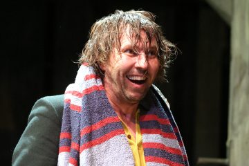 Mies, jolla on sotkuinen, märkä tukka, hymyilee leveästi. Hänellä on raidallinen pyyhe olkapäillään ja keltainen paita, ja hän seisoo tummaa taustaa vasten.