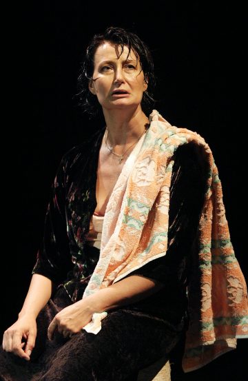 Tummalla taustalla istuu nainen, jolla on märät hiukset, tumma kylpytakki yllään ja olkapäälle ripustettu kukkakuvioinen kylpypyyhe, ja katsoo hieman ylöspäin vakavalla ilmeellä.